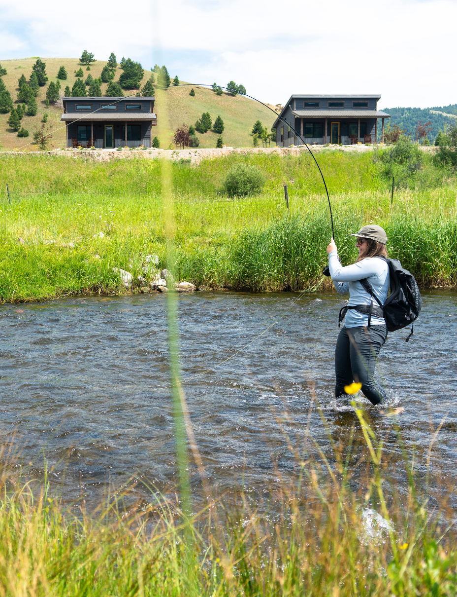 women standing in a small stream holding a bent fly rod in front of cabins