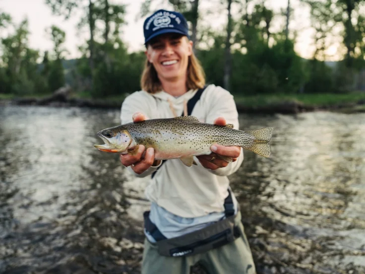 a man holds a cutthroat trout by a river