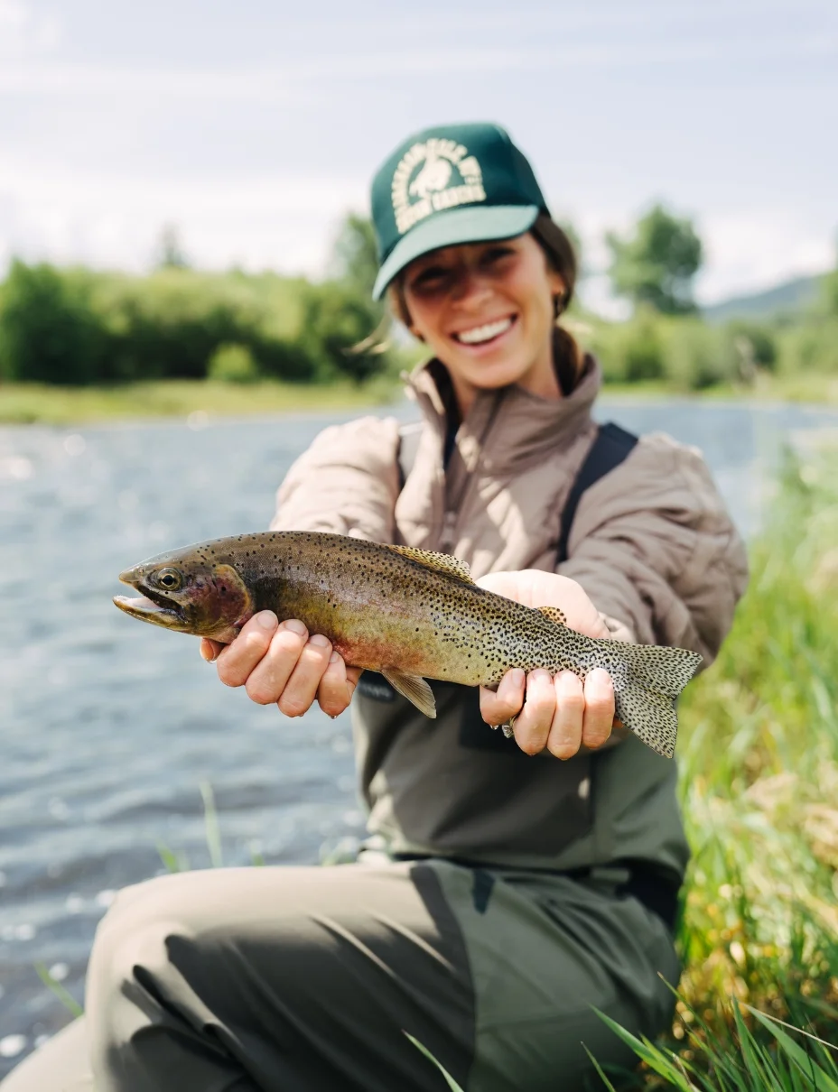 a woman holds a cutthroat trout above a creek