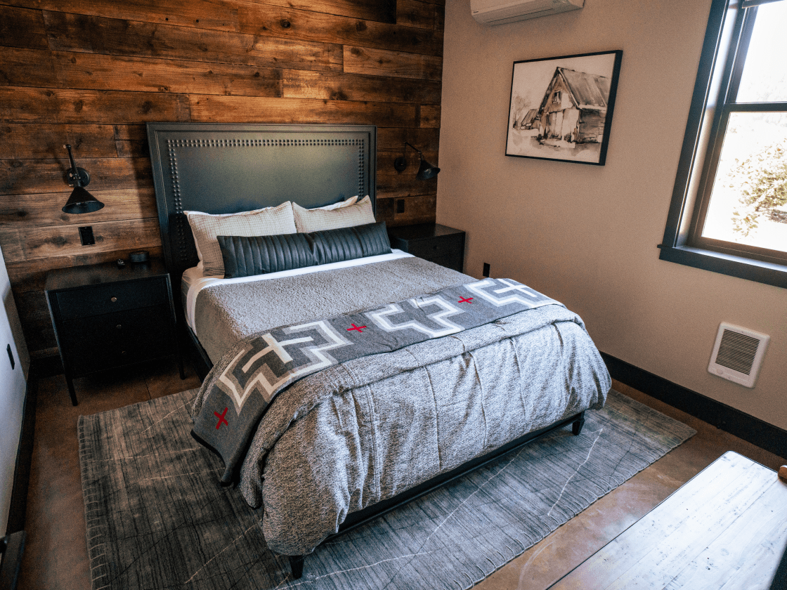 a comfortable bed ina a bedroom with a gray comforter and throw blanket with a pattern. The back wall is a deep colored wood and there is a gray rug on the floor.
