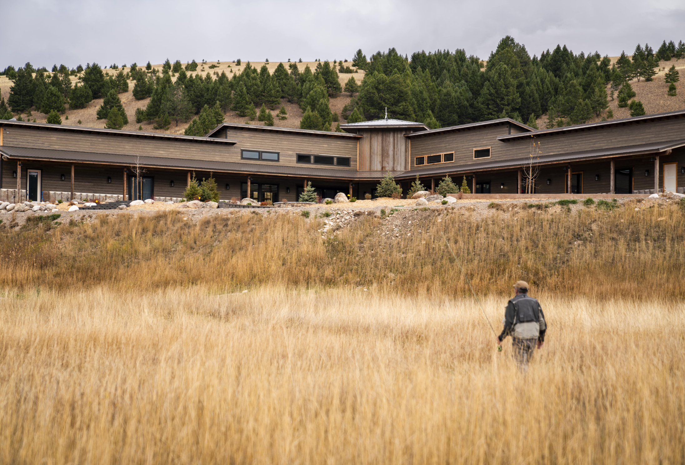 a fly fisherman walks in yellow grass in front of a large brown building