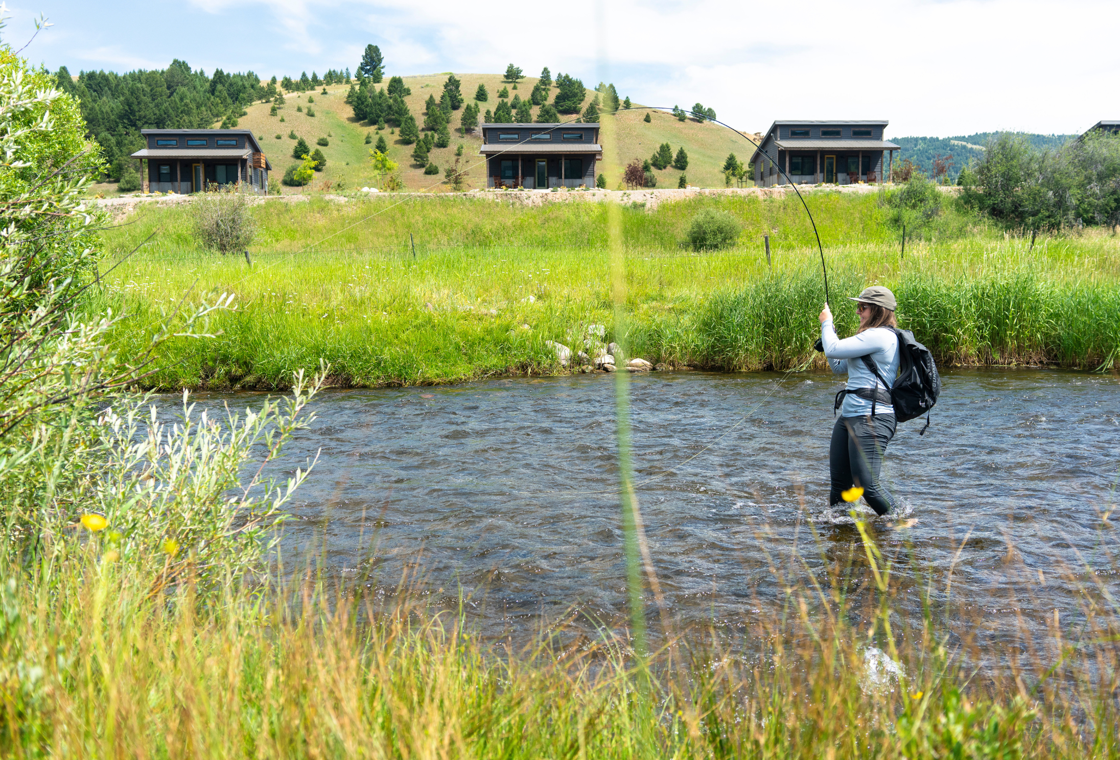 a woman angler stands in a creek fighting a fish on a fly rod in front of cabins