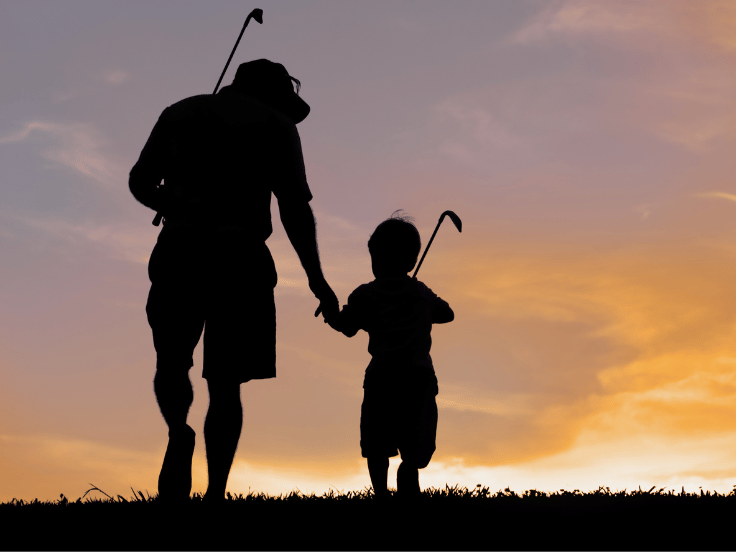 a adult and young child walk with golf clubs over their shoulders at dusk