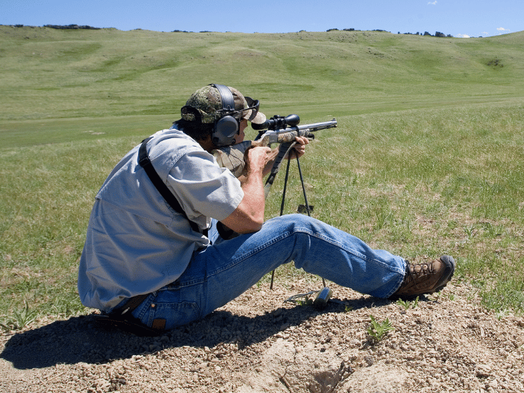 man sits in a grassy field and shoots a rifle