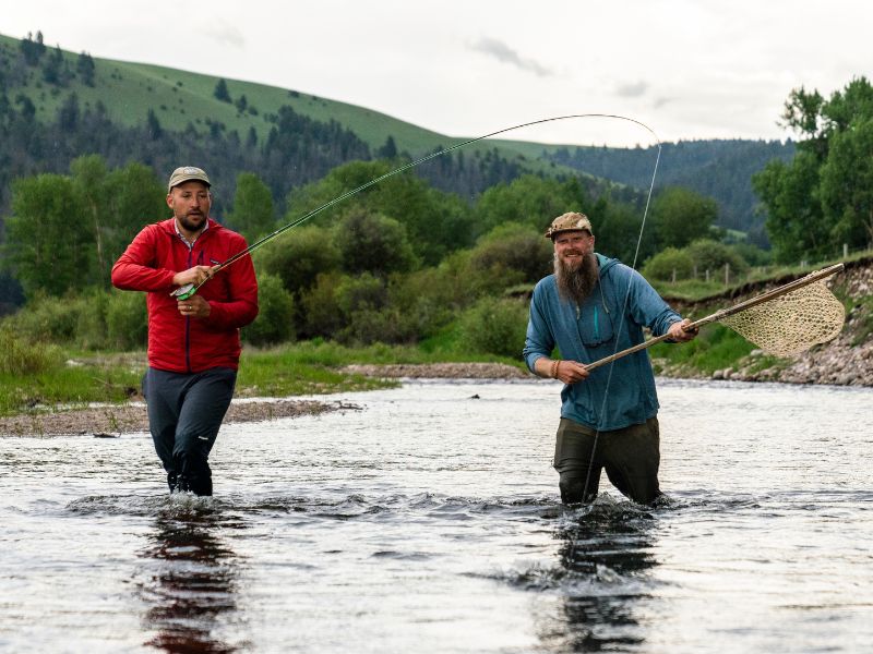 two men stand in a river while they fight a trout on a fly fishing rod