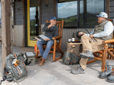 two men sit in chairs in front of a building and talk