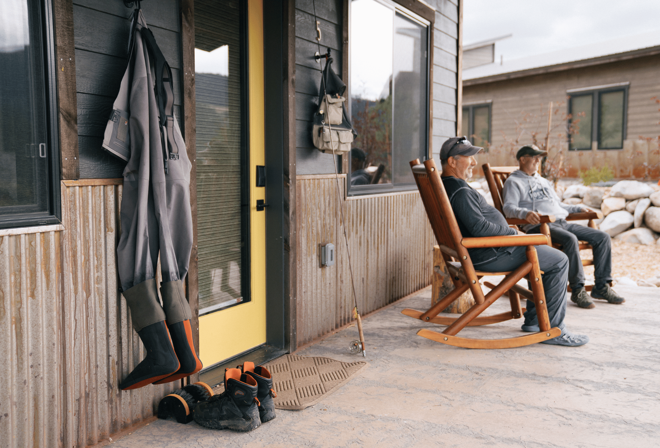 two men sit in rocking chairs by the door of a cabin with a yellow door