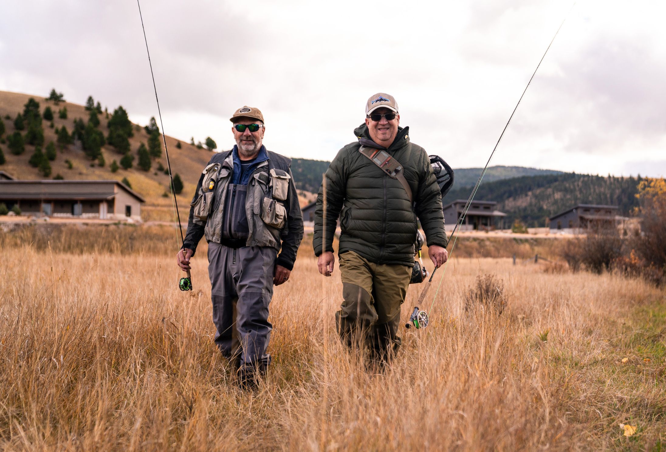 two men walking in a grassy field with fly fishing rods in hand