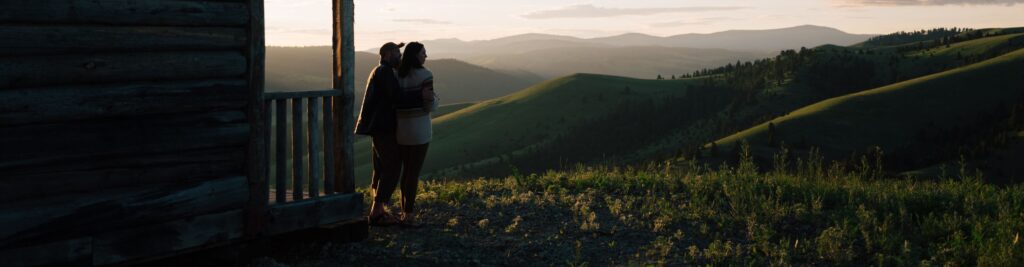 two people standing on top of a hill overlooking the mountains at sunset