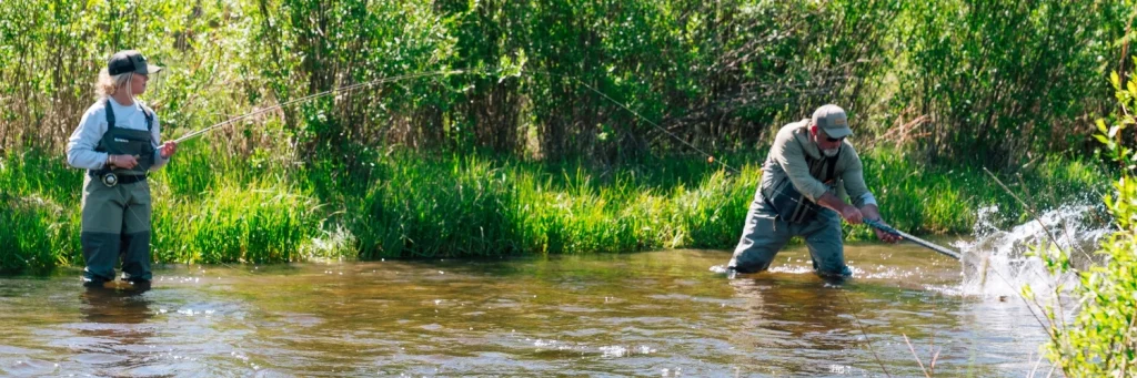 an angler fighting a trout on a fly rod while a guide nets it downstream